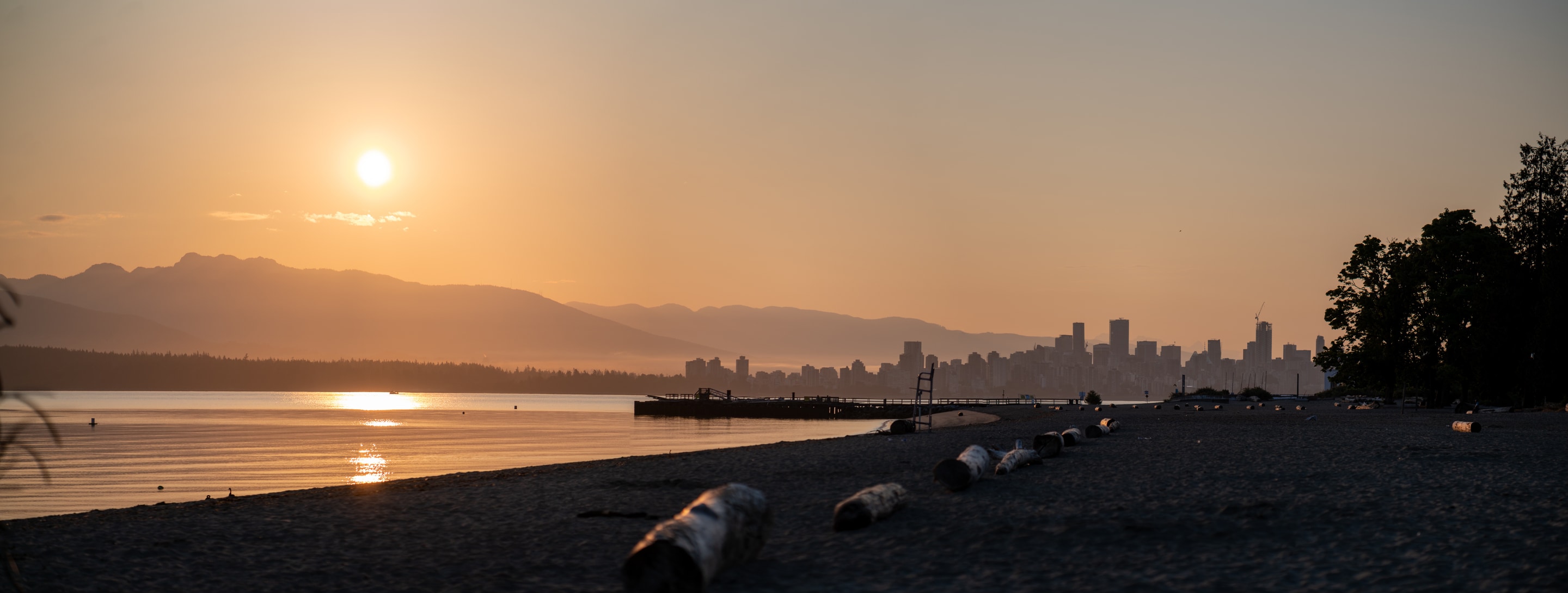 A tranquil scene of a beach at sunset, with the sun setting over the mountains and a city skyline in the distance.