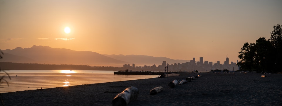 A tranquil scene of a beach at sunset, with the sun setting over the mountains and a city skyline in the distance.