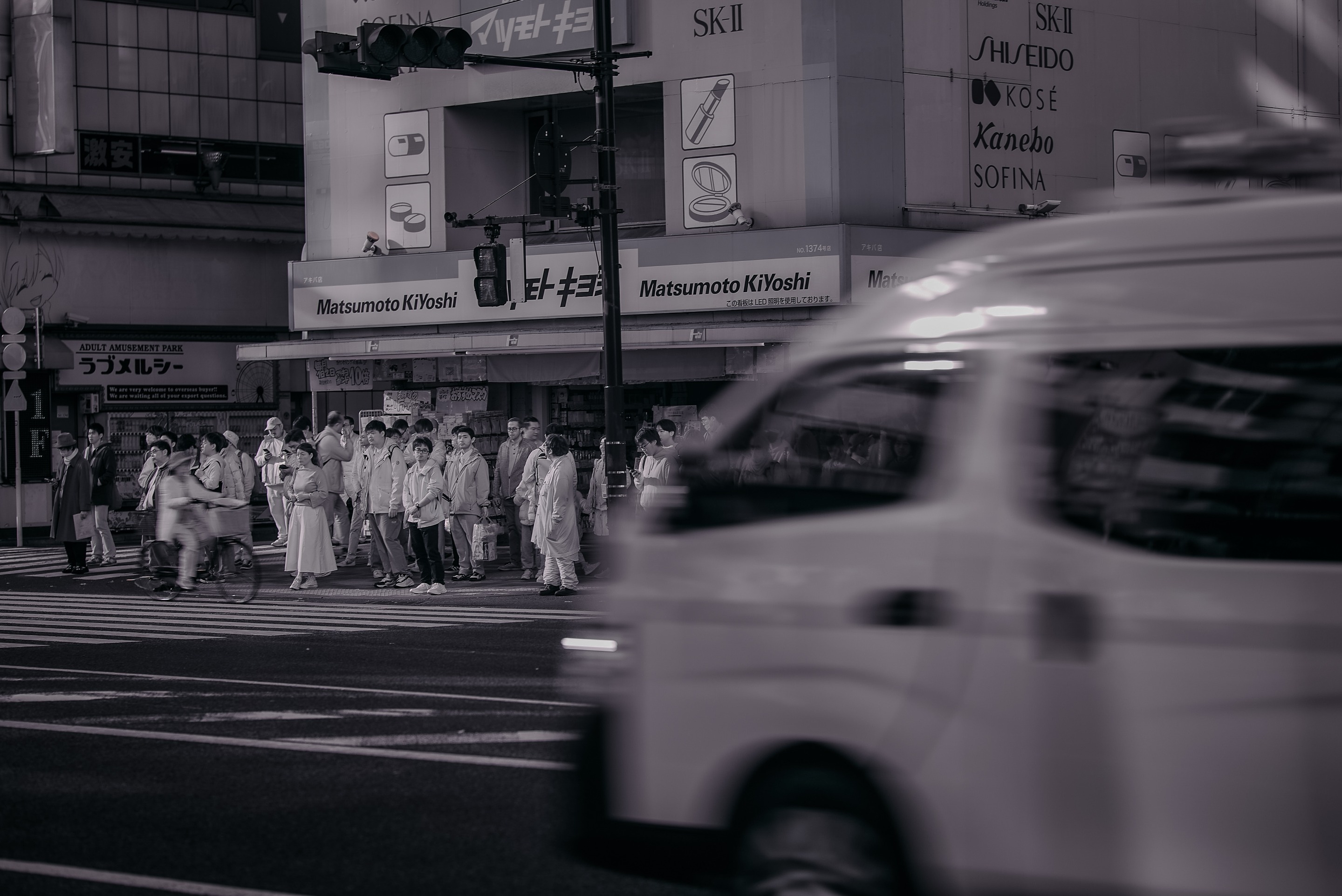 A black and white photograph capturing a busy city street in Japan, with blurred motion from a passing vehicle and a crowd of people waiting to cross at a crosswalk.