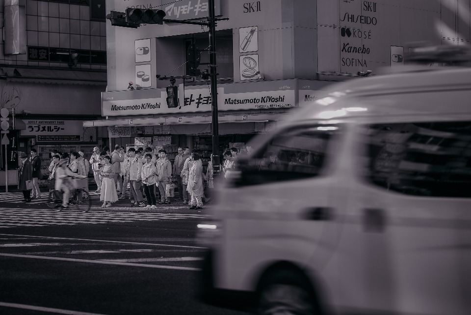 A black and white photograph capturing a busy city street in Japan, with blurred motion from a passing vehicle and a crowd of people waiting to cross at a crosswalk.