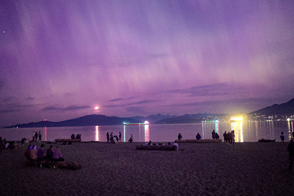 A nighttime beach scene under a vibrant purple sky. Silhouettes of people on the beach contrast with the lights reflecting on the water.