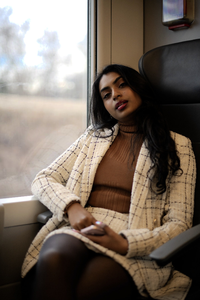 A woman in a blazer and skirt sits on a train, looking at the viewer with a slight smile.