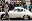 A well-dressed man walks past a vintage cream-colored Porsche. A crowd of people watch the scene. blurred out low res