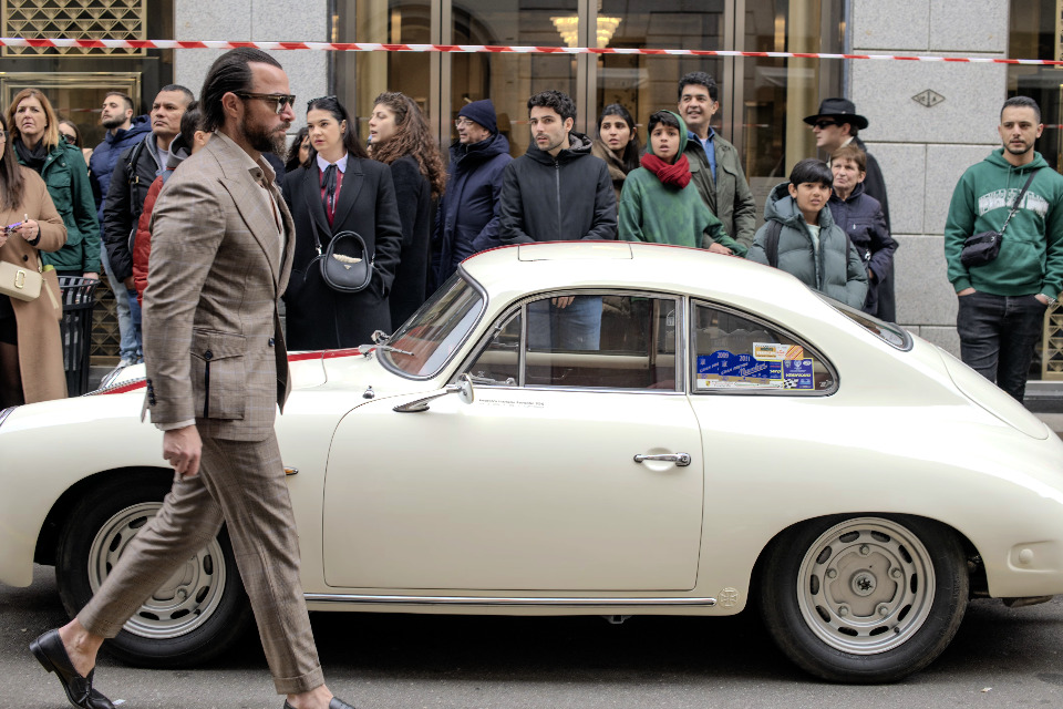 A well-dressed man walks past a vintage cream-colored Porsche. A crowd of people watch the scene.