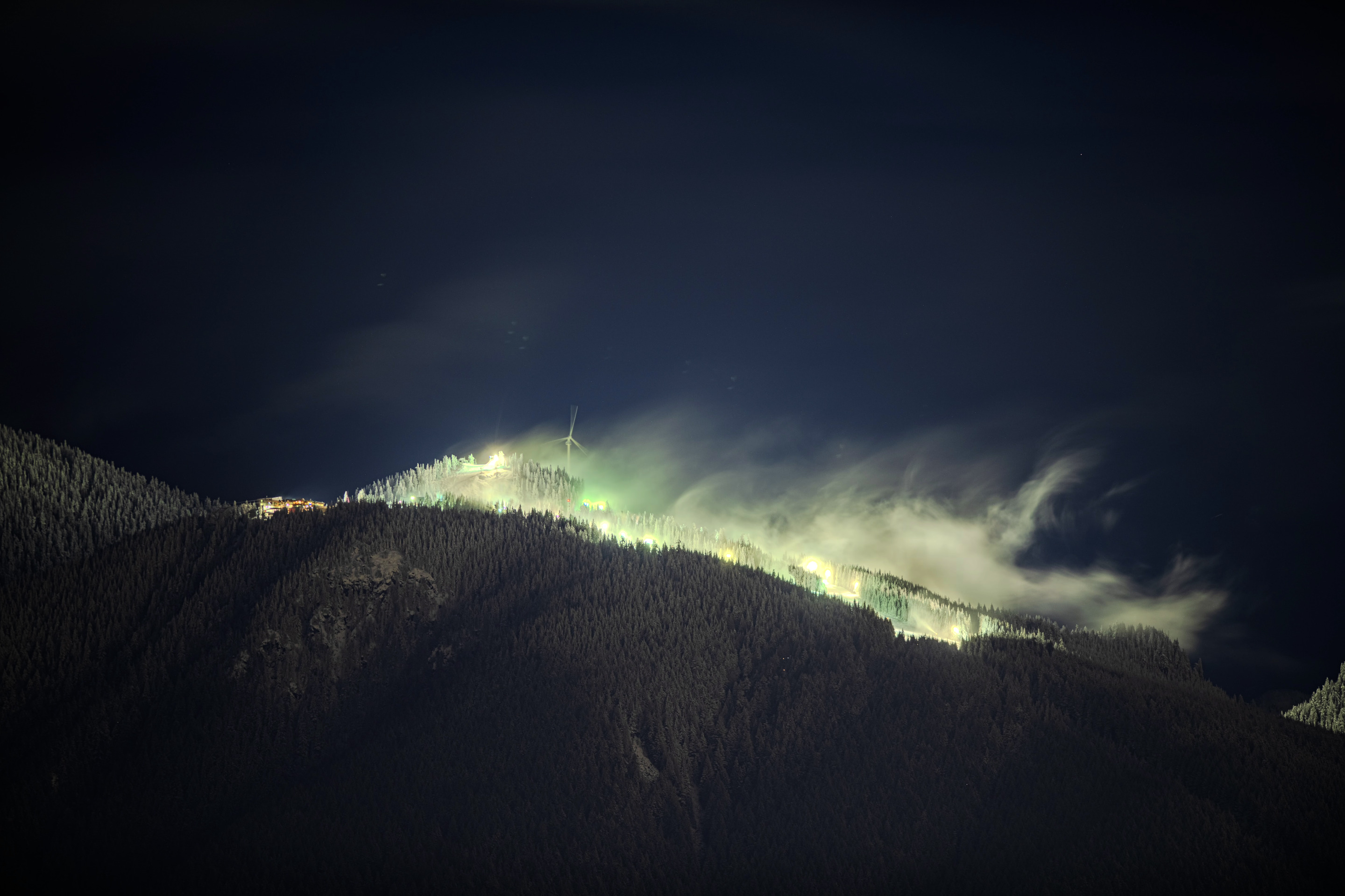 A nighttime landscape shows a mountain range with illuminated slopes and a cloudy sky. The scene is illuminated by lights scattered across the mountain.